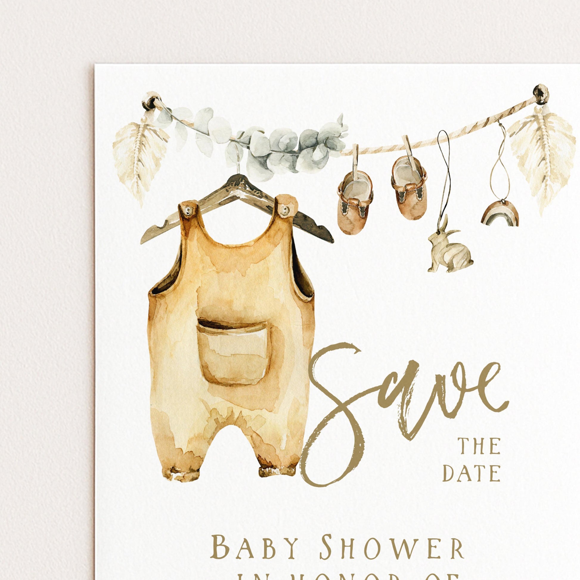 The image is a watercolor painting of a baby's outfit hanging on a hanger, with a pair of shoes and a rabbit nearby. The painting is accompanied by the text "Save the date" and "Baby shower in honor of"