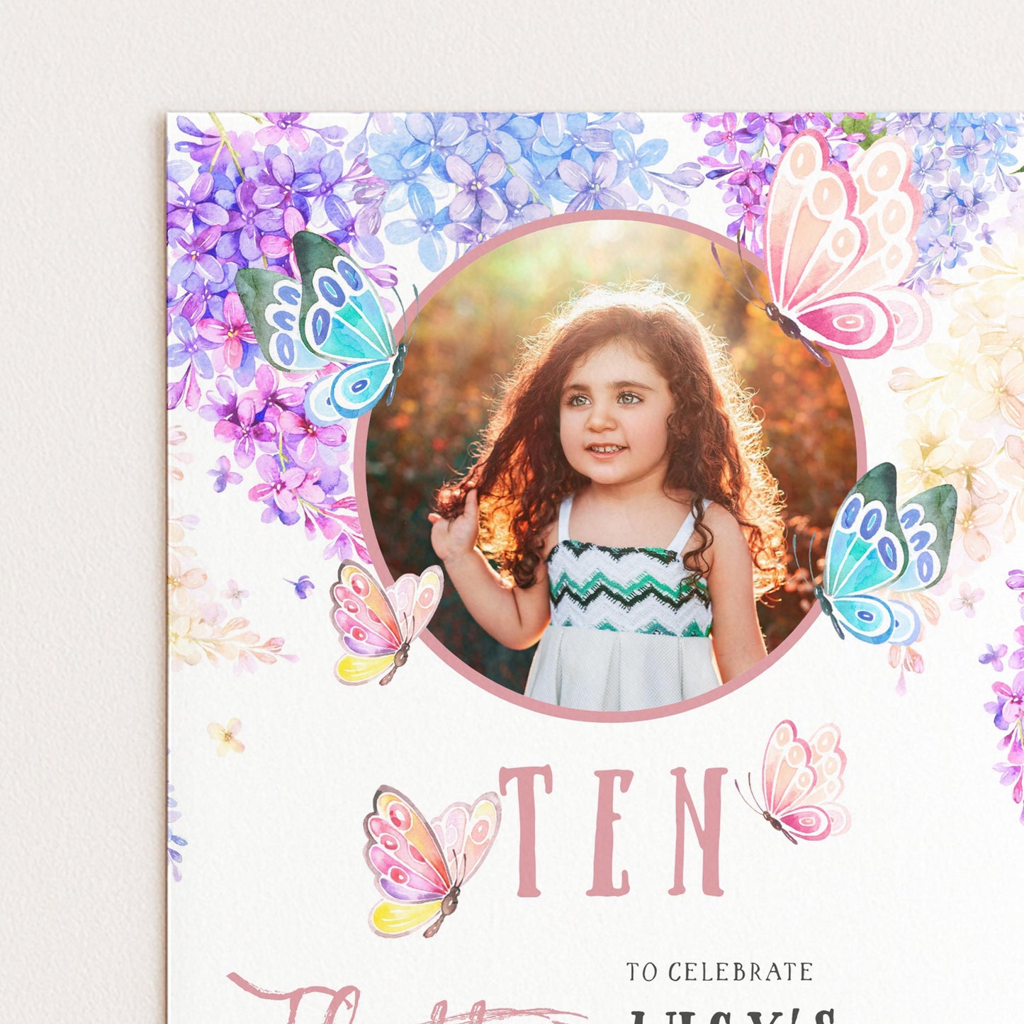 A young girl with curly hair is smiling and posing in front of a floral background with butterflies.