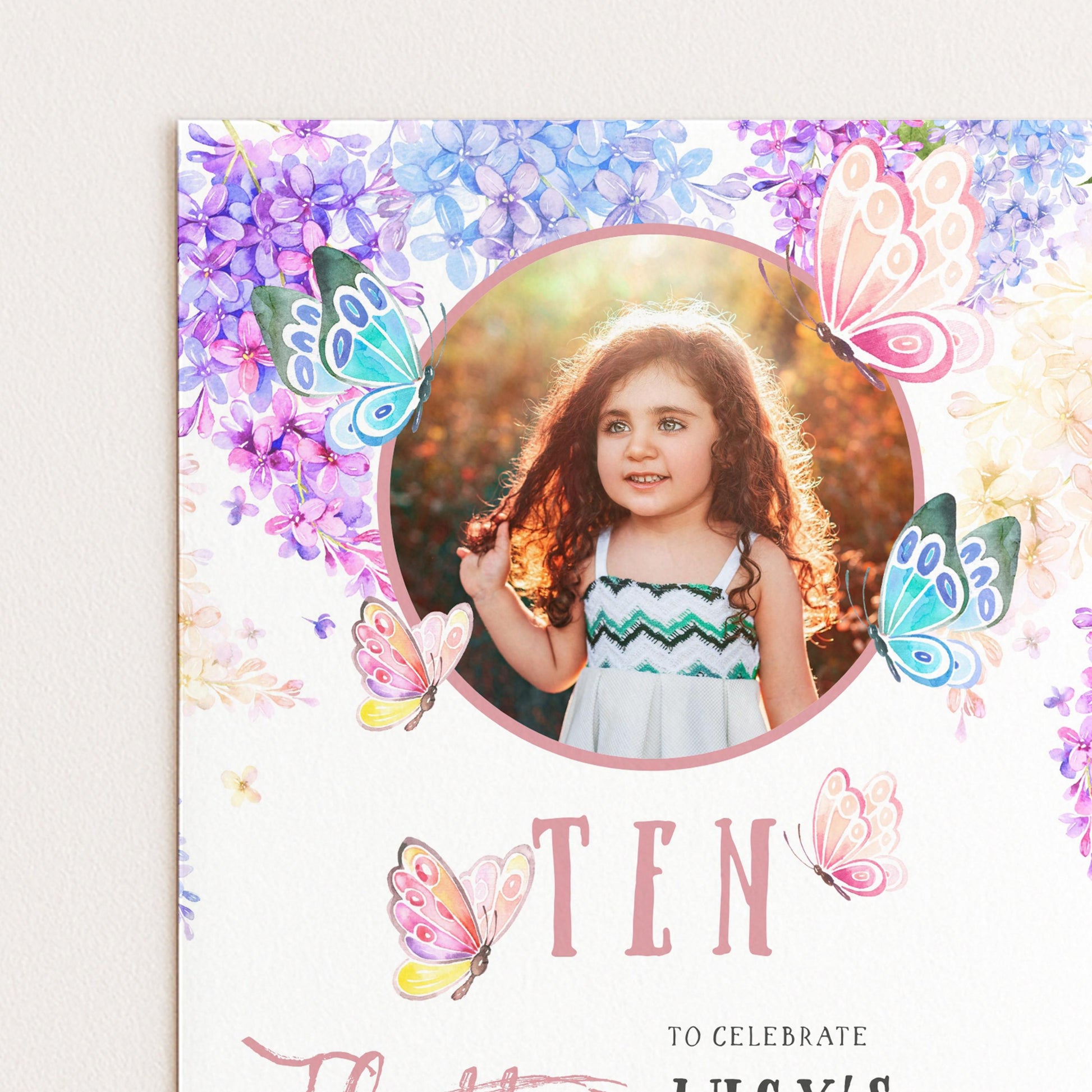 A young girl with curly hair is smiling and posing in front of a floral background with butterflies.