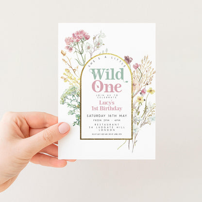 a person holding a white invitation card with a floral design. The card features the text "Wild One" and "Join us to celebrate Lucy's 1st birthday" along with the date and location.