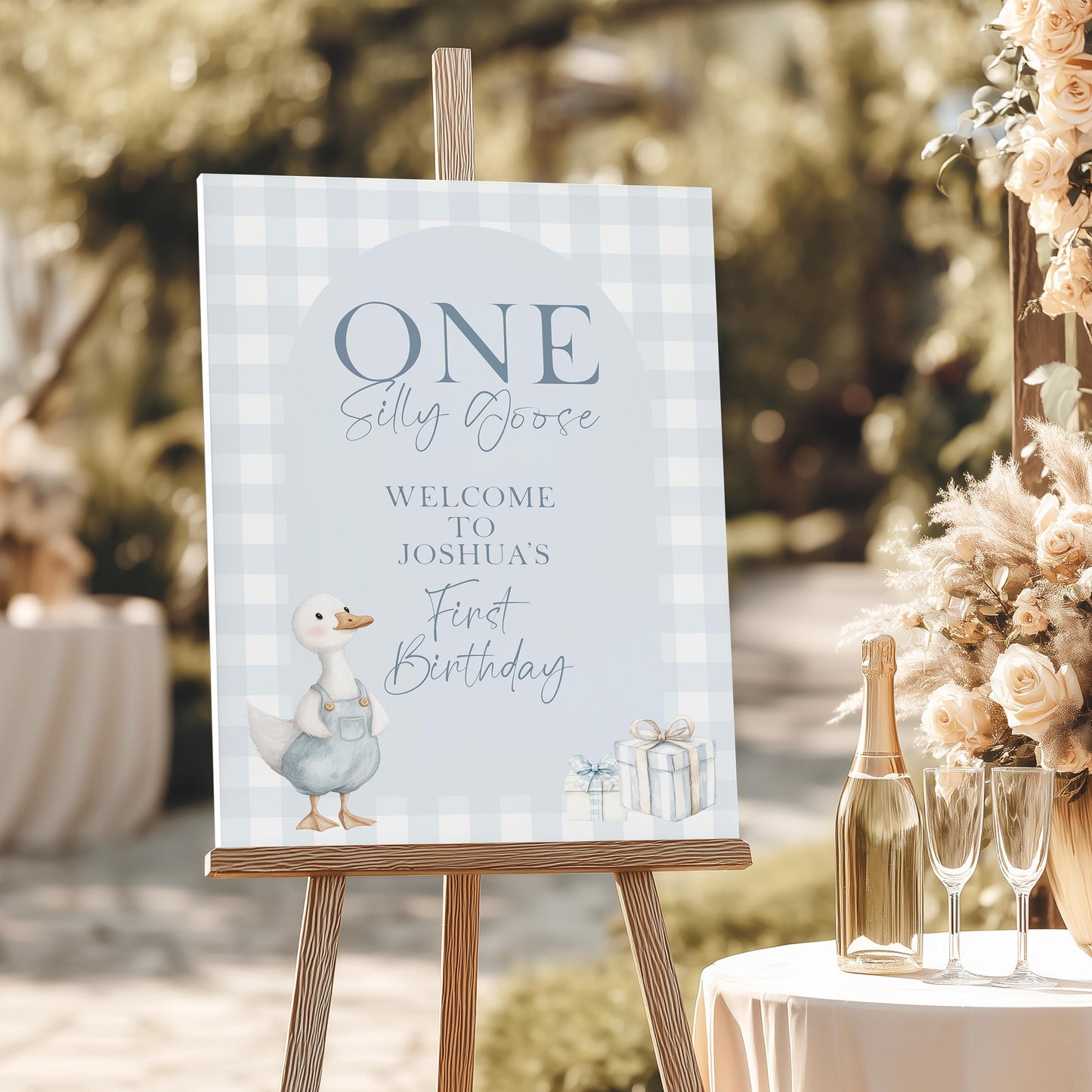 Blue silly goose welcome sign for boy’s 1st birthday, featuring gingham background and illustrated goose in overalls on wooden easel