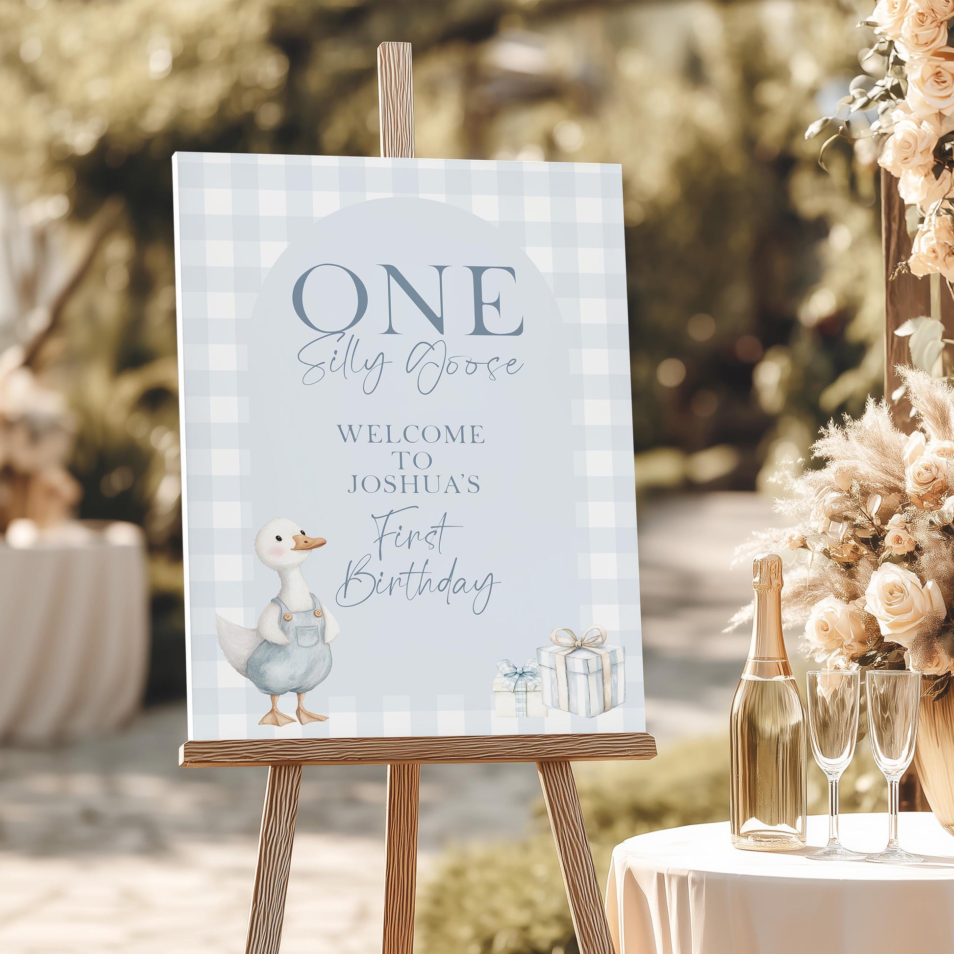 Blue silly goose welcome sign for boy’s 1st birthday, featuring gingham background and illustrated goose in overalls on wooden easel
