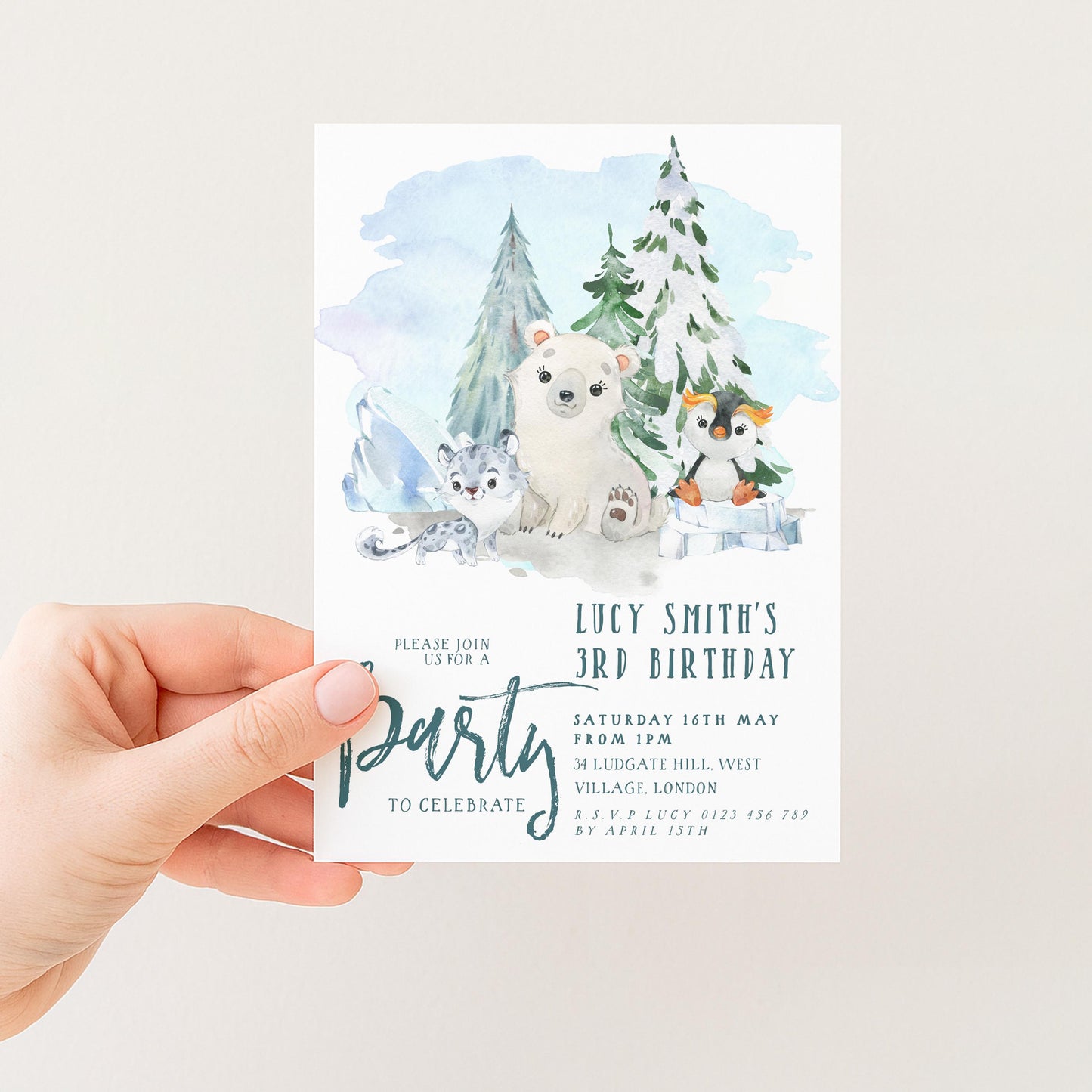 A hand holding a birthday invitation card with a winter scene featuring a polar bear, penguins, and snow-covered trees.