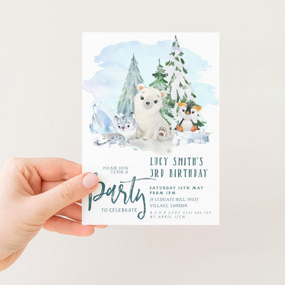 A hand holding a birthday invitation card with a winter scene featuring a polar bear, penguins, and snow-covered trees.
