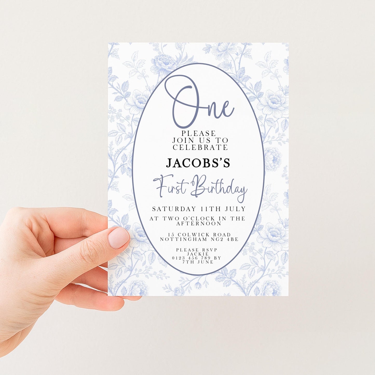 a person holding a white invitation card with a floral design. The card is for a first birthday celebration and includes details about the event, such as the date, time, location, and contact information.