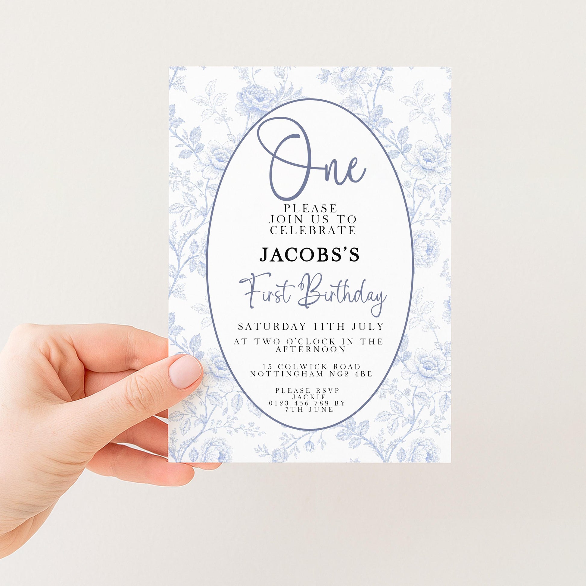 a person holding a white invitation card with a floral design. The card is for a first birthday celebration and includes details about the event, such as the date, time, location, and contact information.