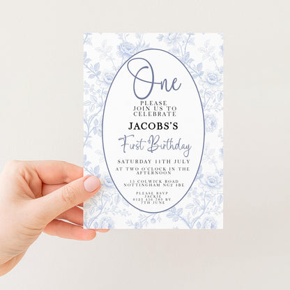 a person holding a white invitation card with a floral design. The card is for a first birthday celebration and includes details about the event, such as the date, time, location, and contact information.