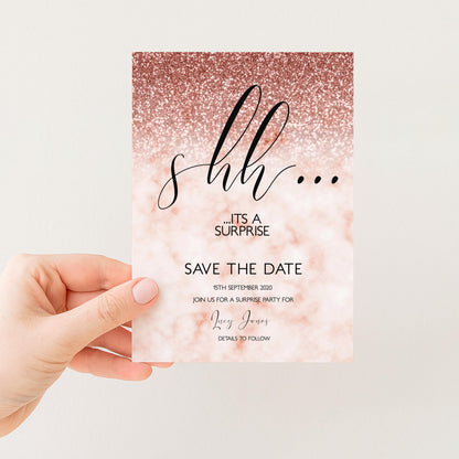 a hand holding a pink and gold glittery invitation card with the text "It's a surprise" and "Save the date" printed on it.