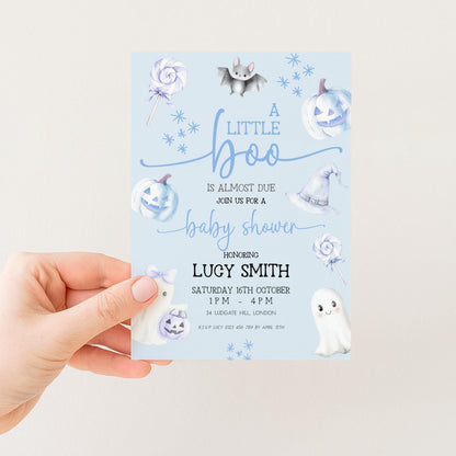 A hand holding a baby shower invitation with a light blue background and various Halloween-themed illustrations, including bats, pumpkins, and ghostly figures.