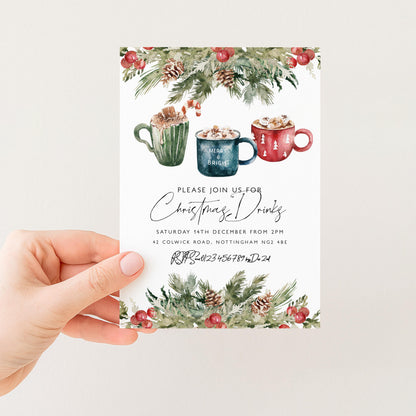 A hand holding a Christmas-themed invitation card with illustrations of mugs and pine branches.