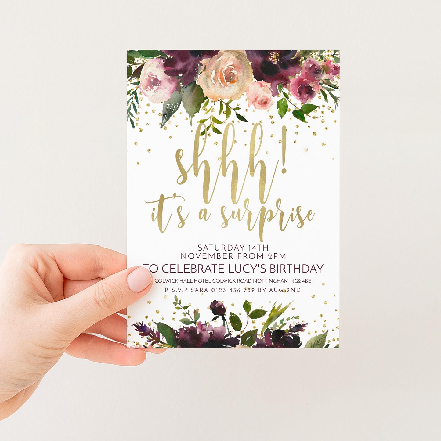 A hand holding a white invitation card with a floral design and gold text.