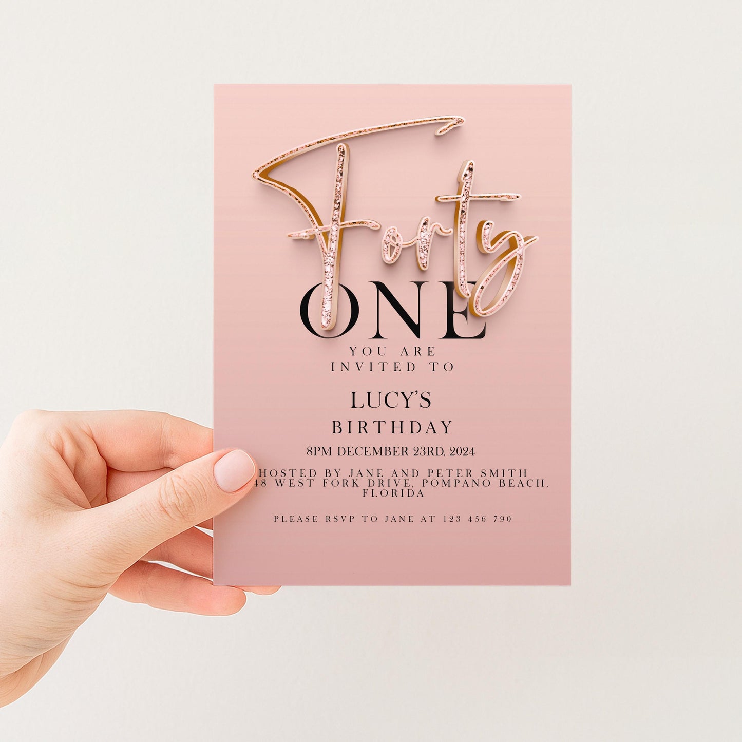 A hand holding a pink invitation card with text and a gold cursive font design.