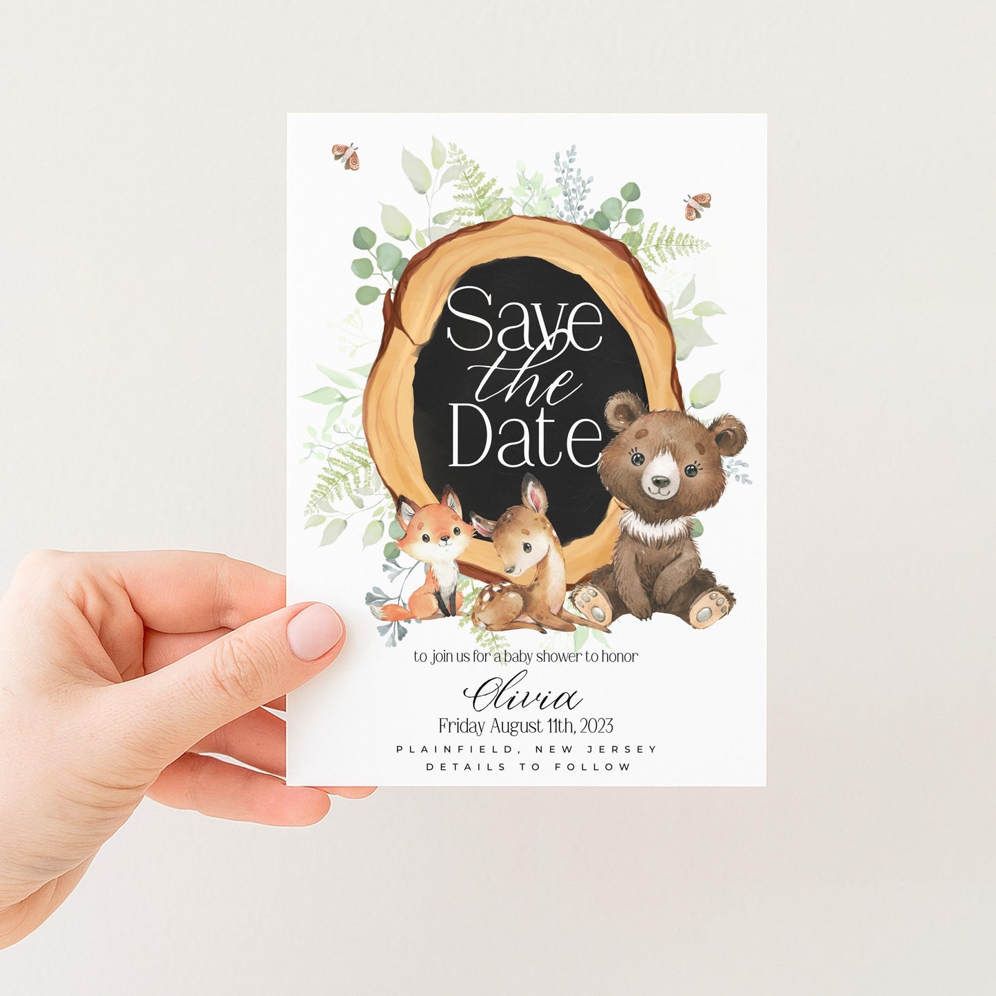 A hand holding a white card with a black border featuring illustrations of a bear, a rabbit, and a deer, along with the text "Save the Date" and a date.