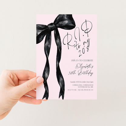 a hand holding a pink invitation card with a black ribbon design.