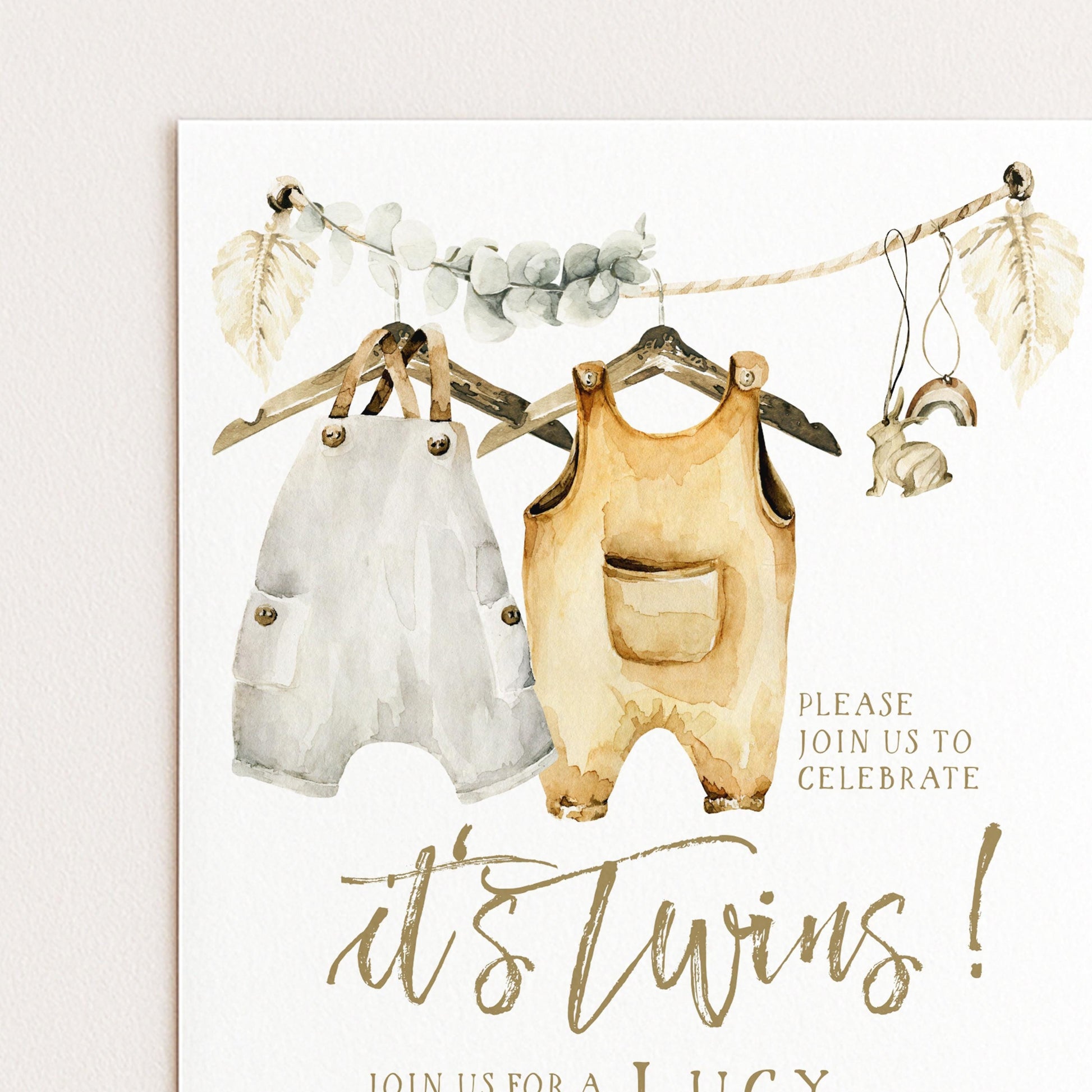 The image is a watercolor painting of baby clothes, including white onesies and yellow jumpsuits, hanging on a clothesline. The painting is accompanied by text inviting guests to a baby shower celebration.