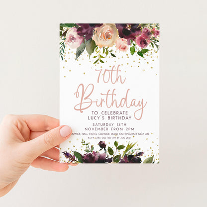 A hand holding a white card with a floral design and text announcing a 70th birthday celebration for Lucy.