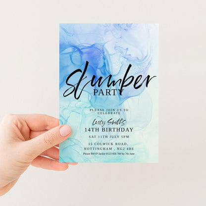 A hand holding a blue and white invitation card for a slumber party.