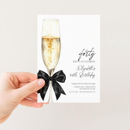 a hand holding a card with a champagne glass and a black ribbon, which appears to be a birthday invitation or celebration card.