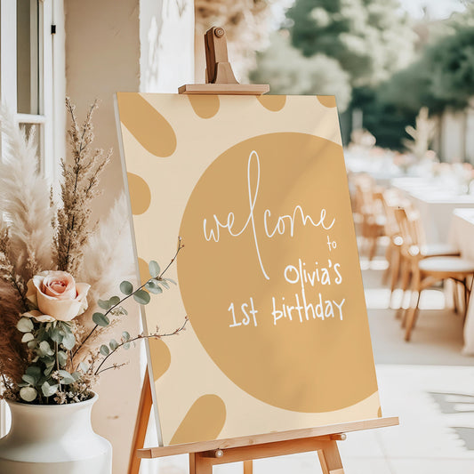 a sign welcoming guests to a first birthday celebration, with a rose in a vase and a table with chairs in the background.