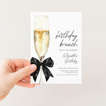 A hand holding a birthday invitation card with a champagne glass and a black bow.