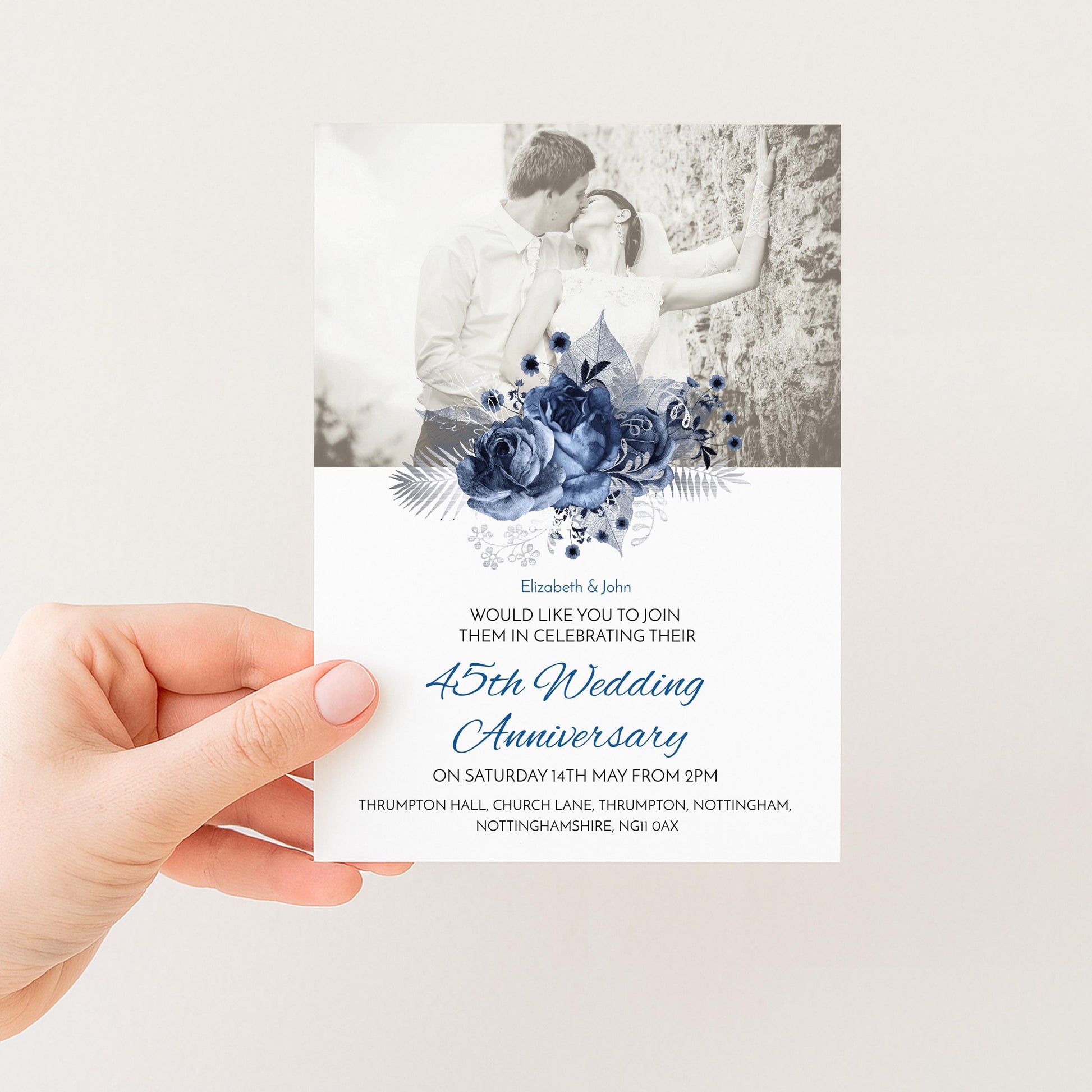 A hand holding a wedding invitation card with a black and white photograph of a couple embracing and a blue floral design.