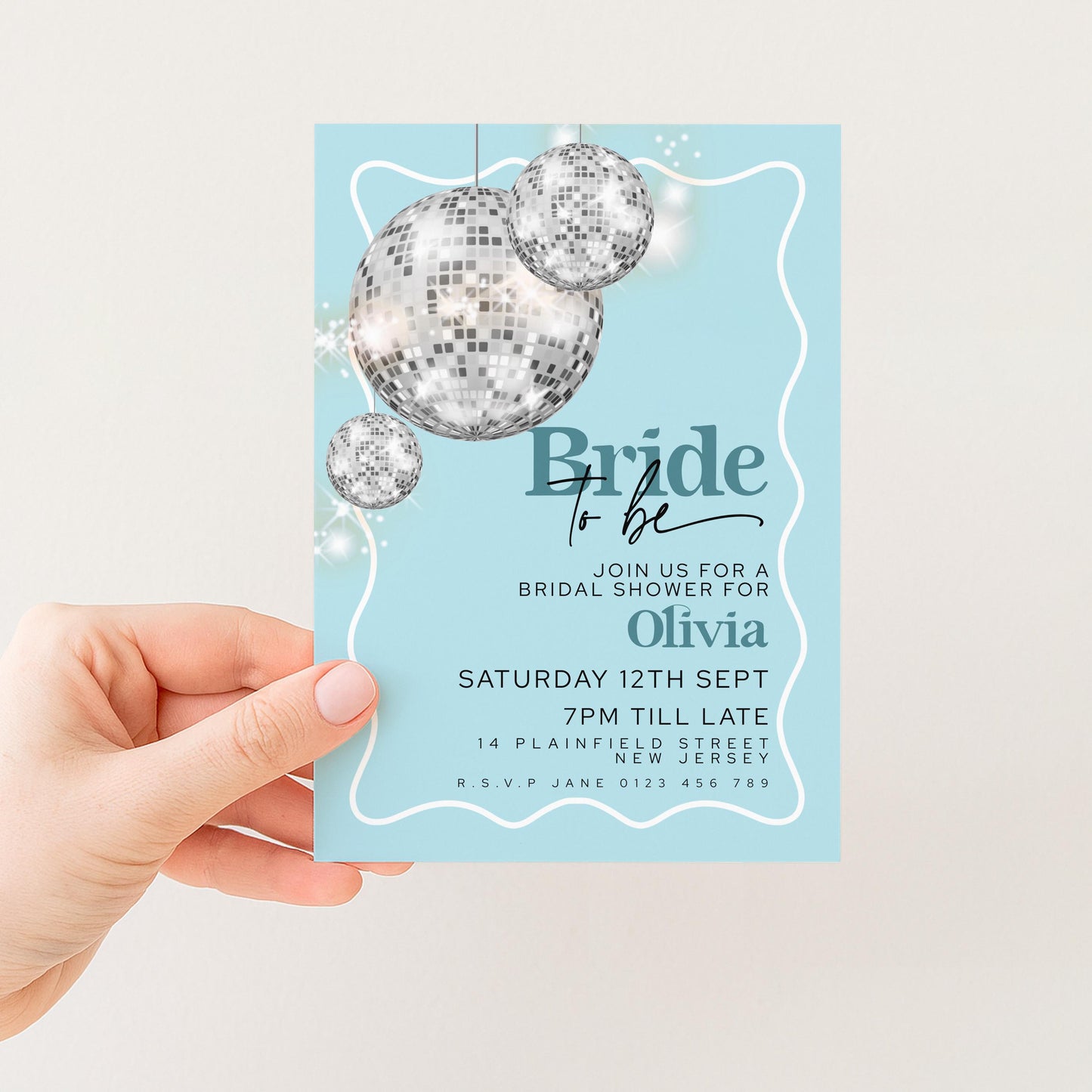 A hand holding a blue invitation card with a disco ball design and text inviting guests to a bridal shower for Olivia.