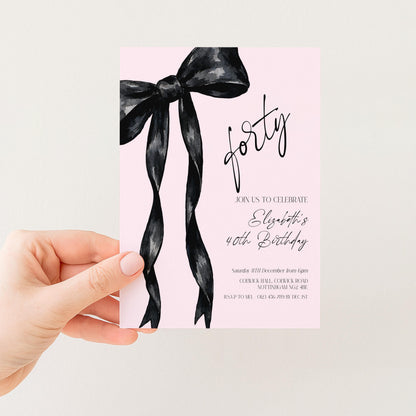 a hand holding a pink invitation card with a black ribbon design.
