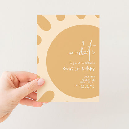 a hand holding a card with a yellow and brown color scheme, featuring a circular design with a sun-like pattern. The card appears to be a save the date card for an event, likely a birthday celebration.