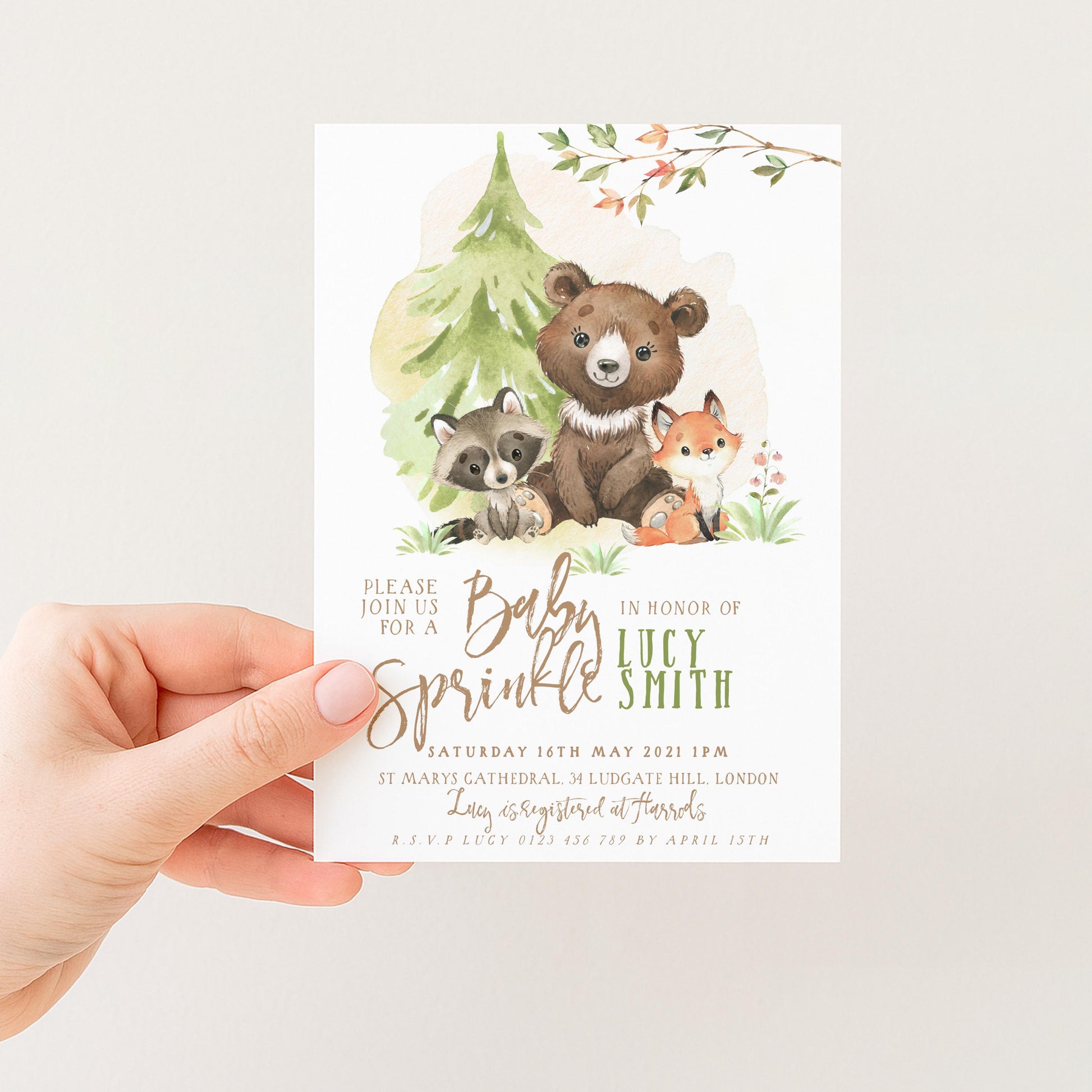 A hand holding a baby shower invitation featuring a bear, raccoon, and fox in a forest setting.