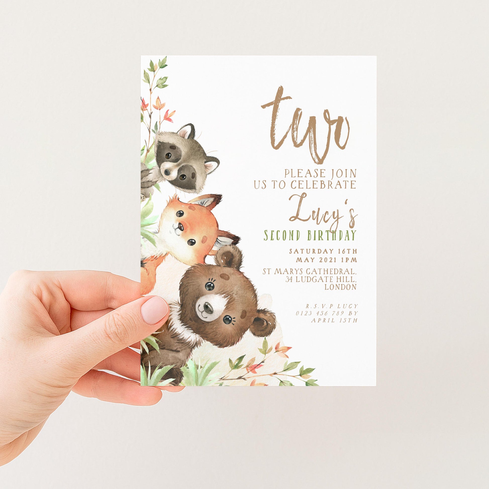 A hand holding a birthday invitation featuring illustrations of a raccoon, a fox, and a bear, along with text inviting guests to a second birthday celebration.