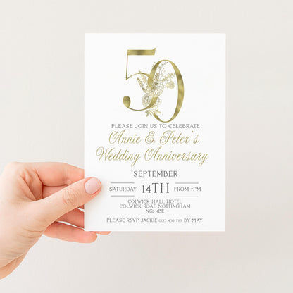 a person holding a white wedding anniversary invitation card with gold lettering and a gold floral design.