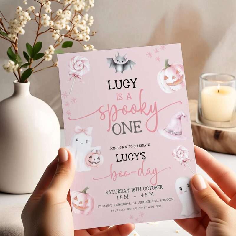 Pastel pink spooky one birthday invitation with cute ghost and pumpkin held in hands, styled with candle and vase decor