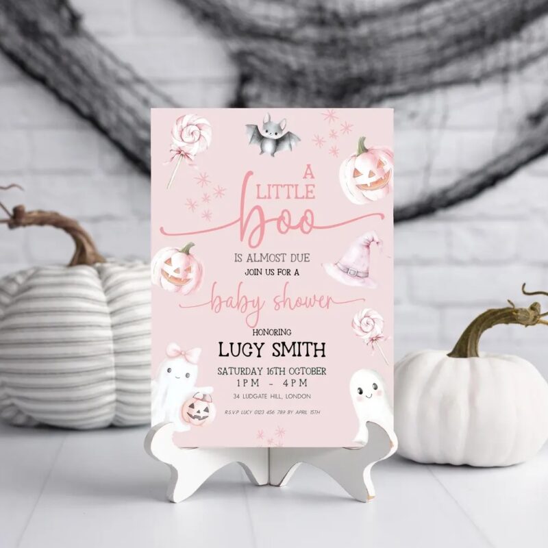 Printed baby shower invitation displayed on stand with white pumpkins and Halloween decor, pink ghost theme with pastel pumpkins