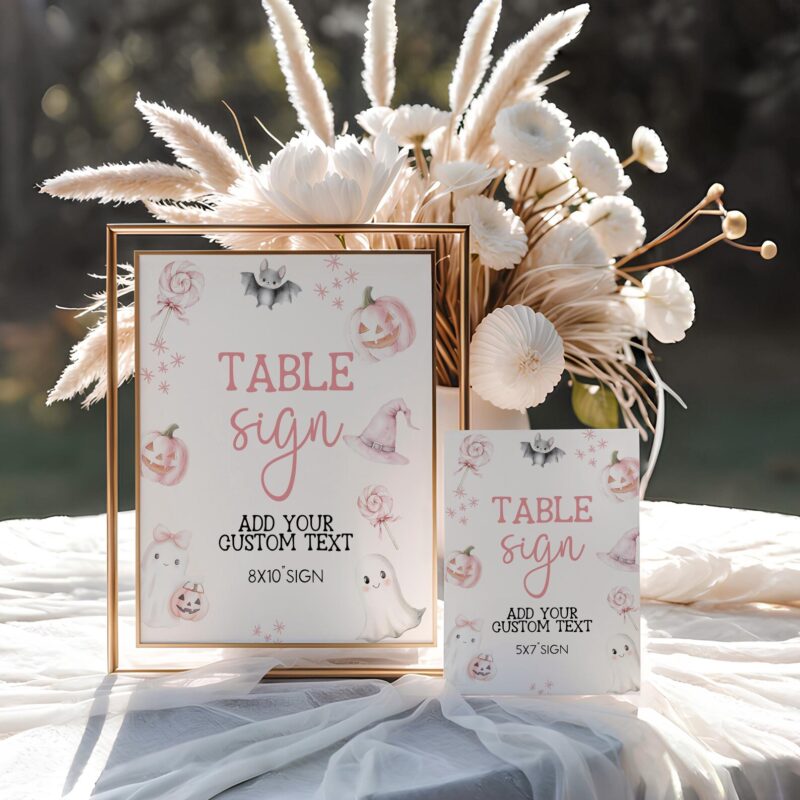 White Halloween baby shower table signs with pink ghosts, pumpkins, and lollipops displayed in gold frames