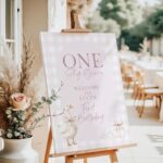 Pink gingham welcome sign for a girl's 1st birthday featuring a goose with a bow and pink gift boxes, styled on an easel with floral party decor.