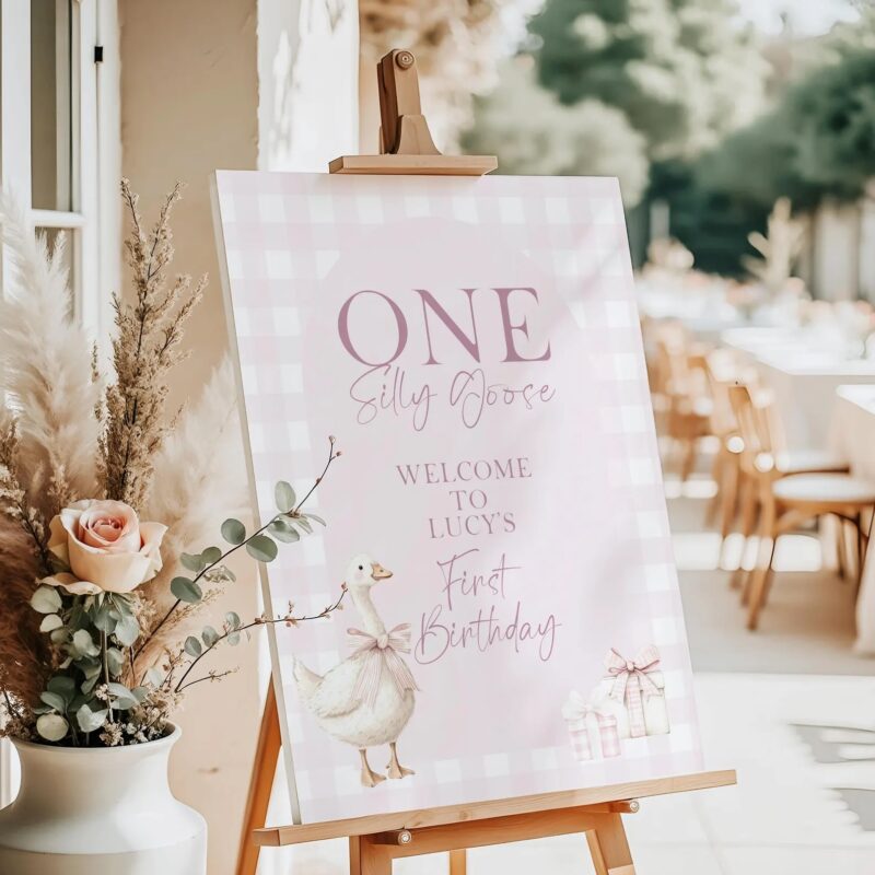 Pink gingham welcome sign for a girl's 1st birthday featuring a goose with a bow and pink gift boxes, styled on an easel with floral party decor.