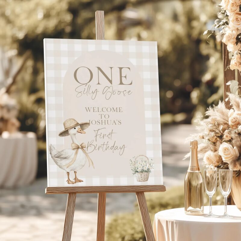 Neutral gingham welcome sign with a hand-drawn goose in a straw hat, displayed on a wooden easel beside champagne and floral decor.